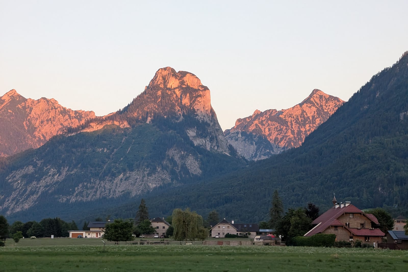 Bergmassiv im Ausseerland, dessen Gipfel im warmen Abendlicht orange leuchten, vor Wiesen und Häusern