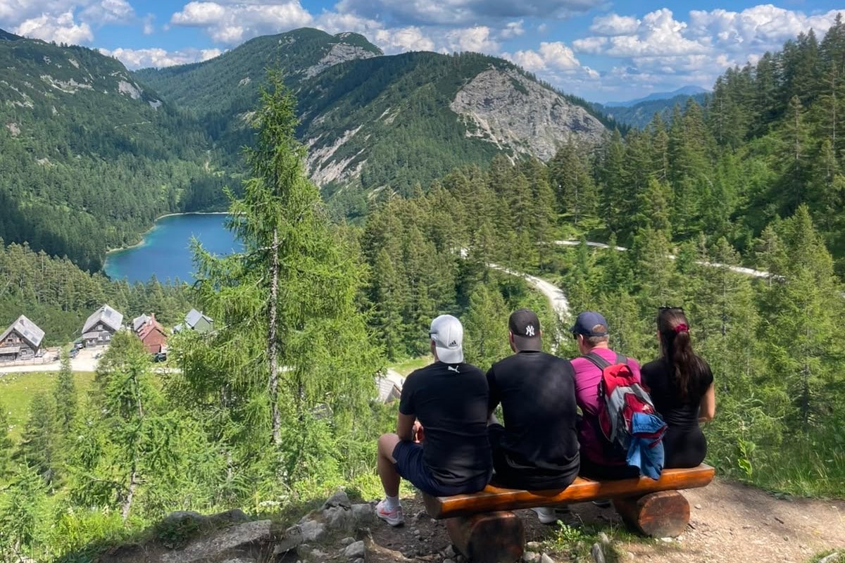 Vier Freunde sitzen auf einer Holzbank über dem blaugrünen Steirersee und blicken in die Berge
