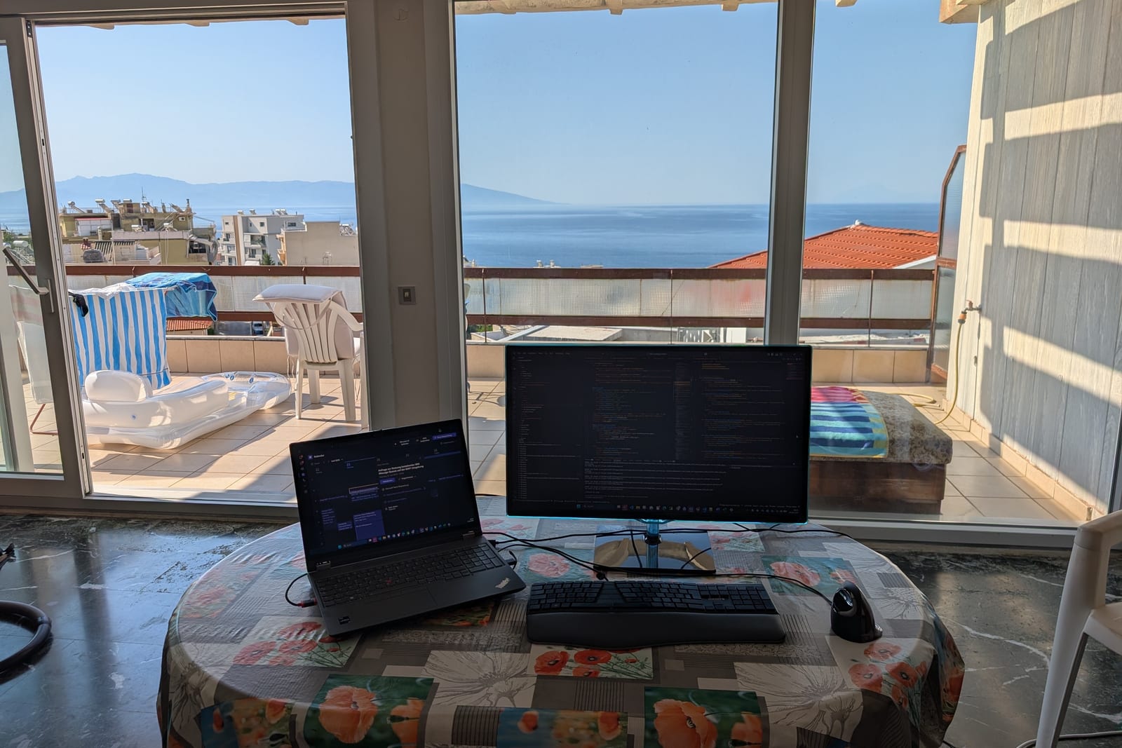 Laptop, großer Monitor, Tastatur und Maus am Esstisch vor offenen Fenstern mit Blick auf das Meer